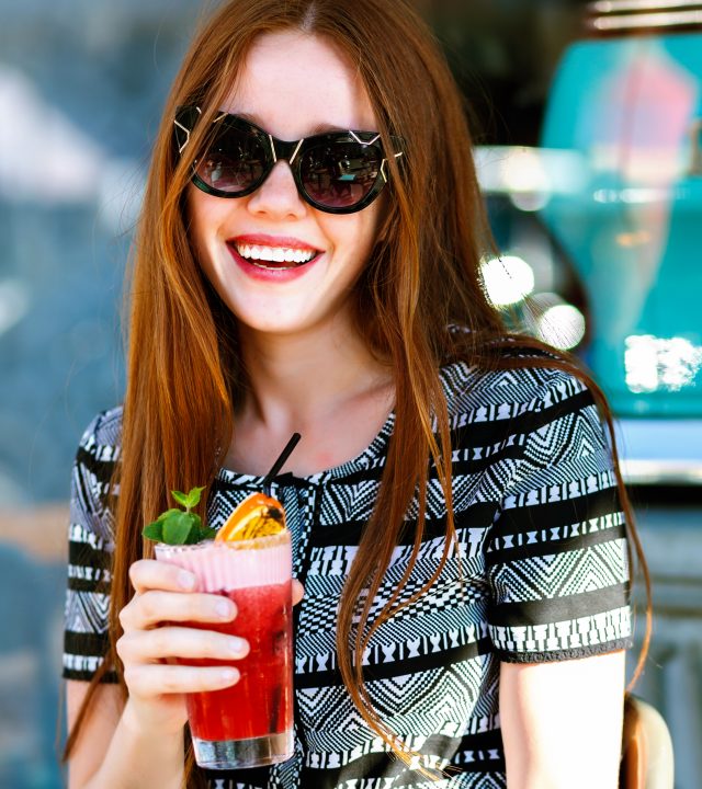 Summer fashion portrait of pretty young elegant ginger woman, sitting at terrace , drinking tasty lemonade, glamour outfit, sunny vacation, relax, joy, natural beauty.